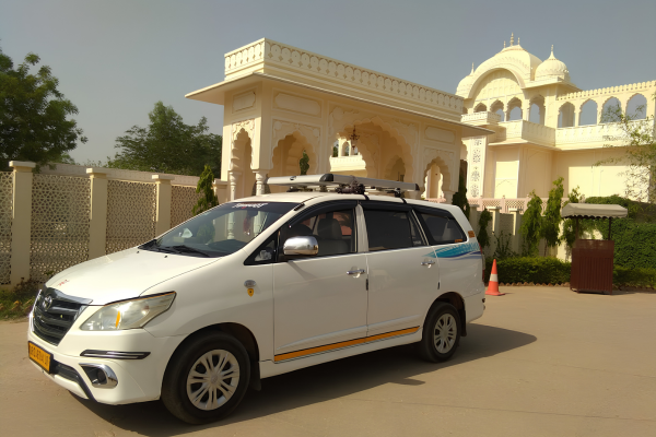 White Toyota Innova parked in front of a heritage hotel and palace-style building in Rajasthan, offering private car rental with driver services.